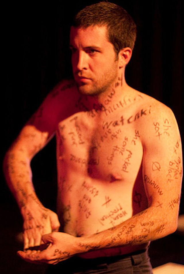 Brandon shirtless and covered in writing, gestures with both hands together