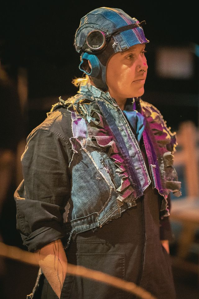 Alison Blakeney in a boiler suit and aviator helmet, with denim ruffles, watching something