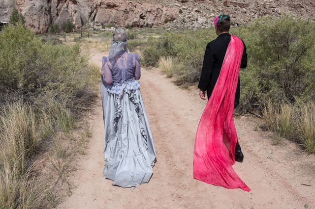 two figures walking away along a desert path, one in a gray dress, the other in a black suit with a pink scarf